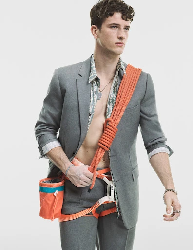 Young man in a suit with climbing gear, including harness, chalk bag, and orange rope slung over his shoulder against a white background.