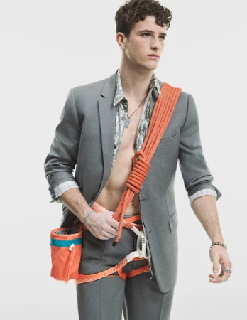 Young man in a suit with climbing gear, including harness, chalk bag, and orange rope slung over his shoulder against a white background.