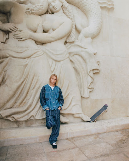 Adult male in a striped outfit leaning against a marble wall with a classical bas-relief sculpture and a skateboard, posing outdoors.