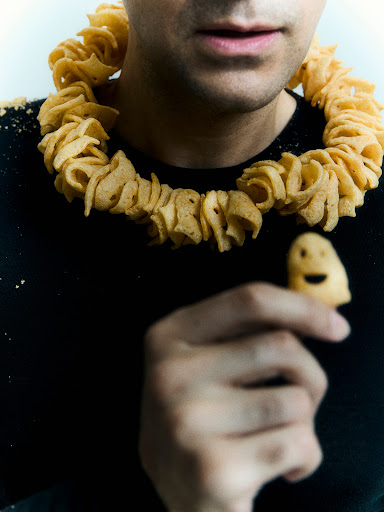 Adult male in a black shirt and potato chip necklace holding a chip with a smiley face, against a plain white background.