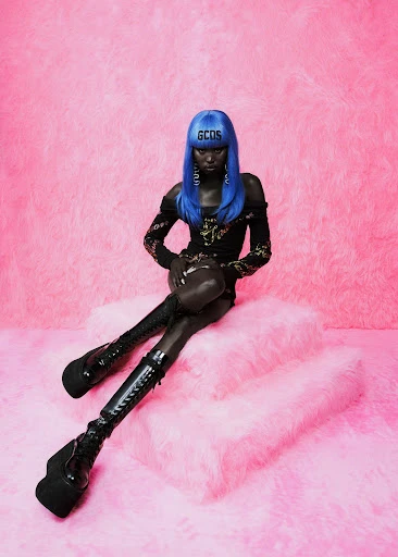 A Black model with blue hair poses on a fluffy platform wearing black boots and a black dress, set against a pink faux fur background.