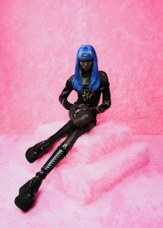 A Black model with blue hair poses on a fluffy platform wearing black boots and a black dress, set against a pink faux fur background.