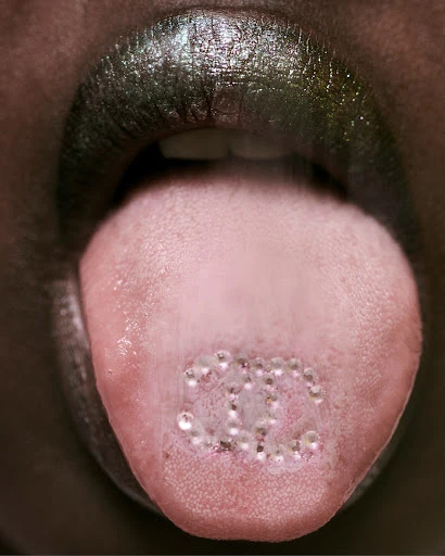 Close-up of an open mouth showing a tongue adorned with small, clear gems, with dark metallic lipstick and a focus on beauty and detail.