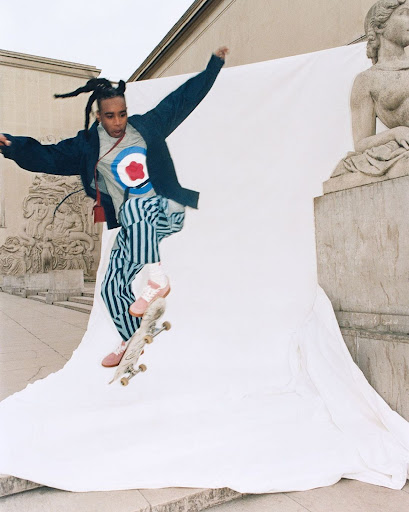 Young adult male jumps on skateboard in an outdoor urban setting with buildings and classical sculpture in the background against a white sheet.