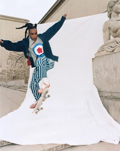 Young adult male jumps on skateboard in an outdoor urban setting with buildings and classical sculpture in the background against a white sheet.