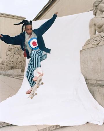 Young adult male jumps on skateboard in an outdoor urban setting with buildings and classical sculpture in the background against a white sheet.