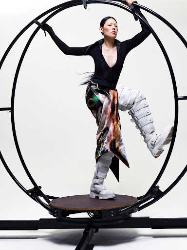 A woman in sculptural boots and a patterned skirt stands on a rotating platform in front of a dark-colored metal wheel apparatus.