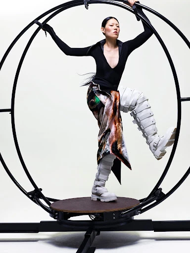 A woman in sculptural boots and a patterned skirt stands on a rotating platform in front of a dark-colored metal wheel apparatus.