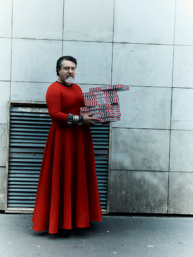 Man in red dress holds stack of pizza boxes against neutral building exterior on city street in a medium, eye-level, full body shot.