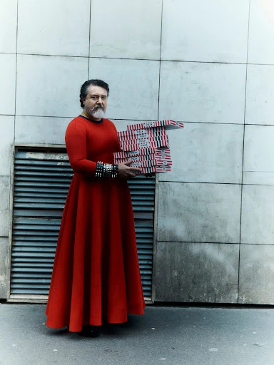 Man in red dress holds stack of pizza boxes against neutral building exterior on city street in a medium, eye-level, full body shot.