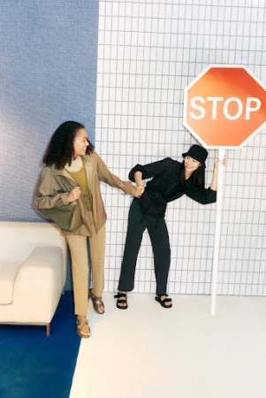 Two women in studio setting: one with a stop sign, another holding her hand. They are wearing casual clothing in an indoor studio.