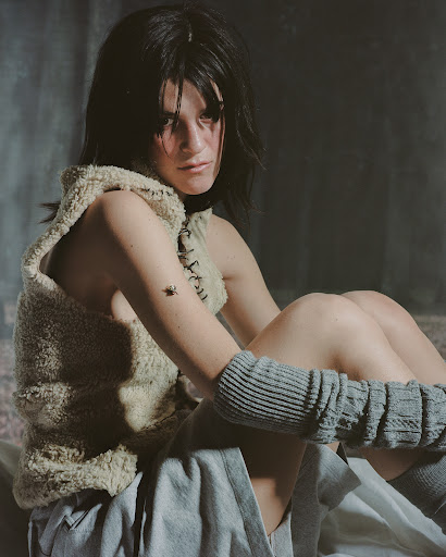 Woman with dark hair sits indoors wearing a sleeveless top, skirt, and knit leg warmers, captured in soft lighting and a shallow depth of field.
