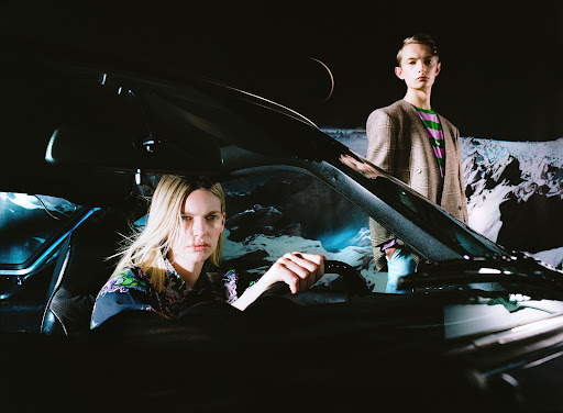 Fashion shot featuring a woman driving a black car and a young male standing outside against a backdrop of a moon and mountains.