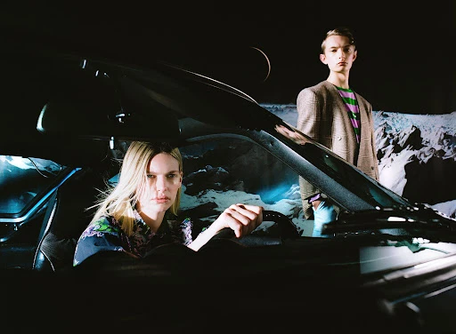 Fashion shot featuring a woman driving a black car and a young male standing outside against a backdrop of a moon and mountains.