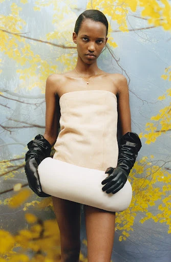 A slender woman poses in a strapless dress and long gloves while holding a pillow, set against a textured background with yellow foliage.
