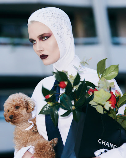 A woman with a white headscarf holds a poodle and a Chanel shopping bag filled with flowers, posing in front of a blurred building.