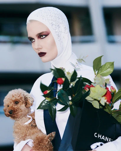 A woman with a white headscarf holds a poodle and a Chanel shopping bag filled with flowers, posing in front of a blurred building.