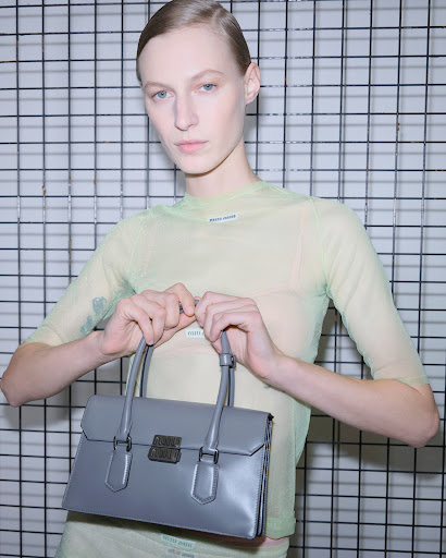 Woman posing with a gray leather handbag in front of a wire rack in a studio with soft, neutral lighting; fashion editorial.