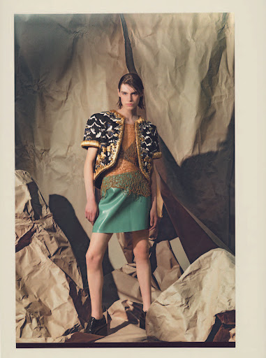 Fashion model in an embellished jacket and skirt poses indoors in front of crumpled paper for a studio portrait.