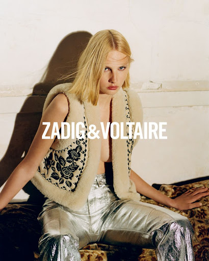 A fair-skinned model with blonde hair poses indoors, wearing a shearling vest and metallic silver pants, showcasing a fashion-forward style.