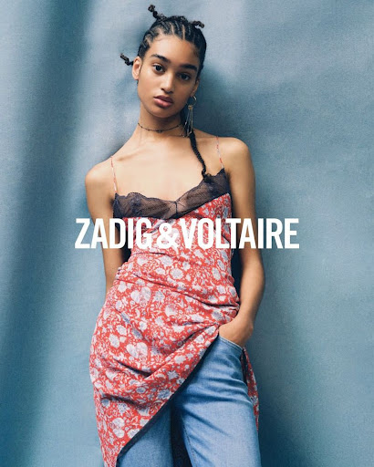 A woman poses in a red floral dress and blue jeans, hands in pockets, against a textured blue background, showcasing a Zadig & Voltaire ad.
