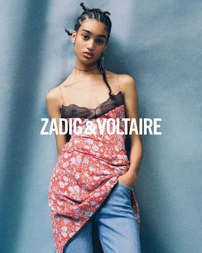 A woman poses in a red floral dress and blue jeans, hands in pockets, against a textured blue background, showcasing a Zadig & Voltaire ad.