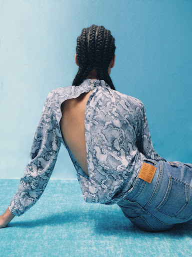Back view of a woman in a snakeskin-patterned top and jeans sitting against a blue backdrop, showcasing fashion and style.