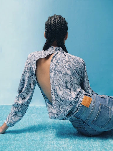 Back view of a woman in a snakeskin-patterned top and jeans sitting against a blue backdrop, showcasing fashion and style.