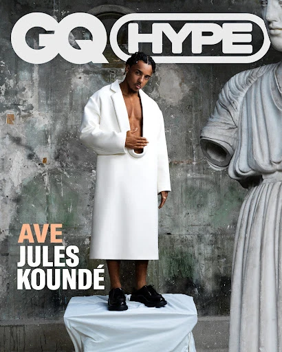 Jules Koundé poses in a white coat with open front, black shoes on a white platform in front of a weathered wall and sculpture.