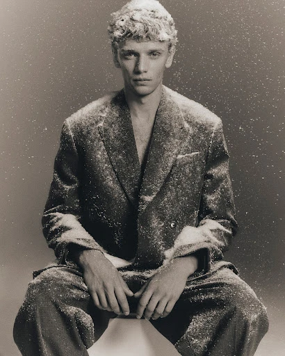 Monochrome portrait of a fair-skinned adult male model in a double-breasted suit covered in white specks, seated on a white surface.
