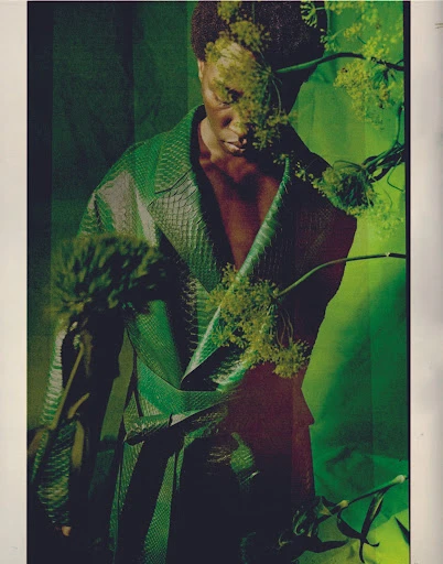 A person in a green snake-skin coat is partially obscured by flowering dill plants against a green backdrop in a studio-style fashion shot.