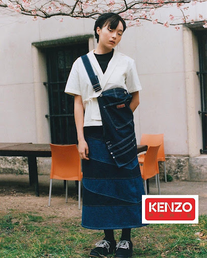 Woman in an ivory shirt and patchwork denim skirt with the KENZO logo. Outdoor fashion shot with a building and trees in the background.
