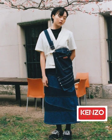 Woman in an ivory shirt and patchwork denim skirt with the KENZO logo. Outdoor fashion shot with a building and trees in the background.