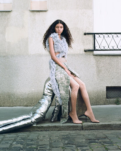 Fashion photography: Young woman poses outdoors wearing a grey feather top, camouflage skirt, fishnet stockings, and high-heel shoes.