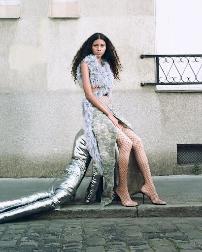 Fashion photography: Young woman poses outdoors wearing a grey feather top, camouflage skirt, fishnet stockings, and high-heel shoes.