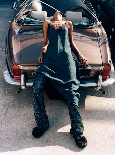 A woman in a dress and pants leans against the trunk of a vintage convertible car in a fashion-style photograph shot at eye level.