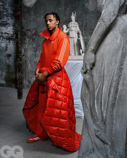 Portrait of adult male fashion model wearing an orange tracksuit and puffer blanket, posing in a studio with stone statues in the background.