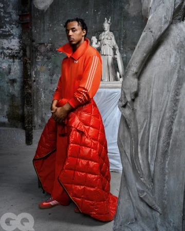 Portrait of adult male fashion model wearing an orange tracksuit and puffer blanket, posing in a studio with stone statues in the background.