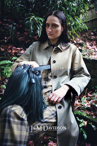 A fashionable woman in a trench coat poses outdoors with a fringed bucket bag, styled in a natural setting with lush green foliage.