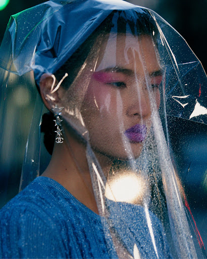Woman with magenta makeup and sculptural plastic head covering, wearing a blue top and Chanel earrings against a dark background.
