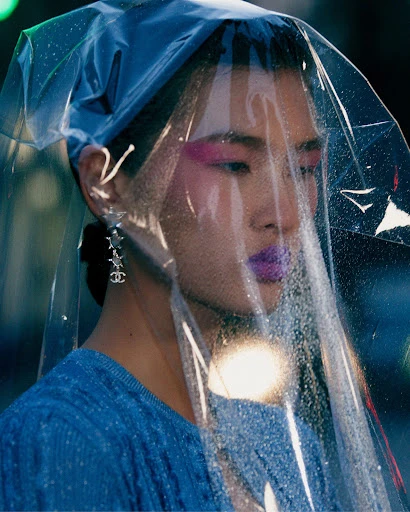 Woman with magenta makeup and sculptural plastic head covering, wearing a blue top and Chanel earrings against a dark background.