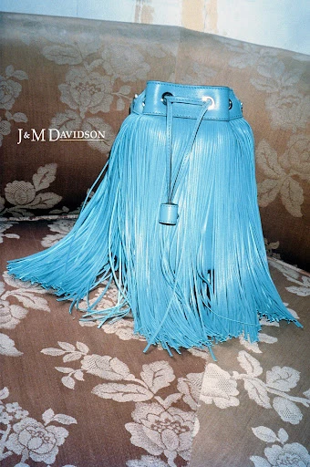 Turquoise fringe bucket bag by J&M Davidson on a floral patterned sofa, displayed in a fashion product photography studio setting.