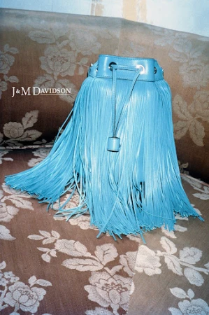Turquoise fringe bucket bag by J&M Davidson on a floral patterned sofa, displayed in a fashion product photography studio setting.