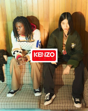 Two models seated on a plaid sofa against a wood-paneled wall, wearing casual sweaters, dark trousers, and sneakers.