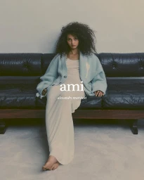 Model with dark curly hair seated on a dark leather sofa wearing an oversized light blue cardigan and cream dress