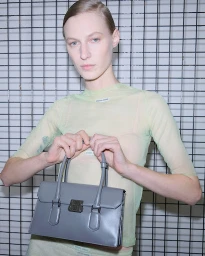 Model in sheer pale green top holding a grey structured leather handbag in front of a metal grid wall.