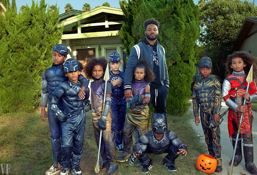 Group of children dressed in superhero costumes with an adult on a grassy lawn in front of a light green house on a sunny day.
