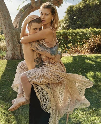 Outdoor portrait featuring a female celebrity in a dress piggybacking on a male celebrity, with green foliage in the background.