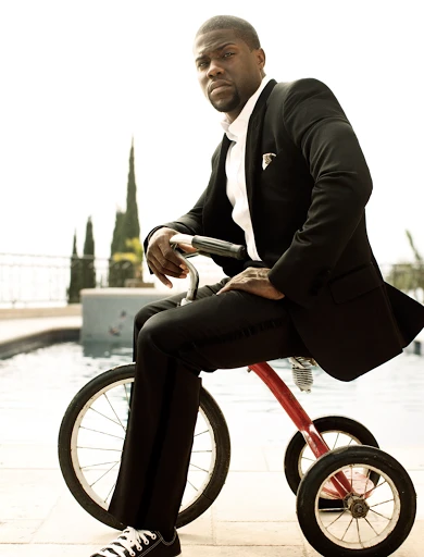 Man in a suit riding a red tricycle outdoors, portrait style with pool and greenery in the background, looking towards the camera.