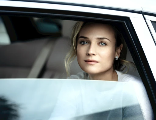 Close-up portrait of Diane Kruger looking through the open window of a car, wearing a white shirt and diamond earrings.